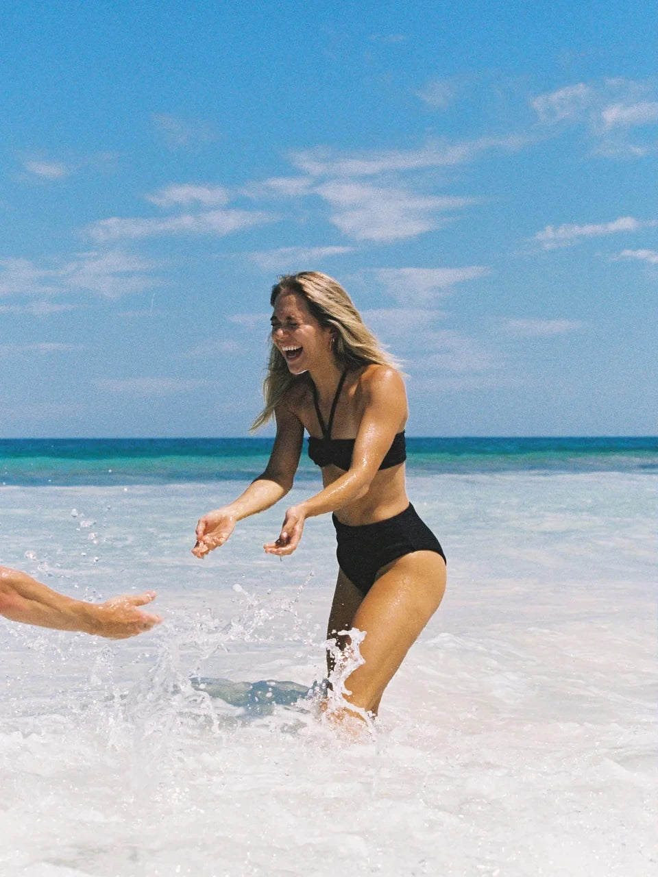 Layday Towels, A Woman in a black bikini standing in shallow water with a clear blue sky and ocean background