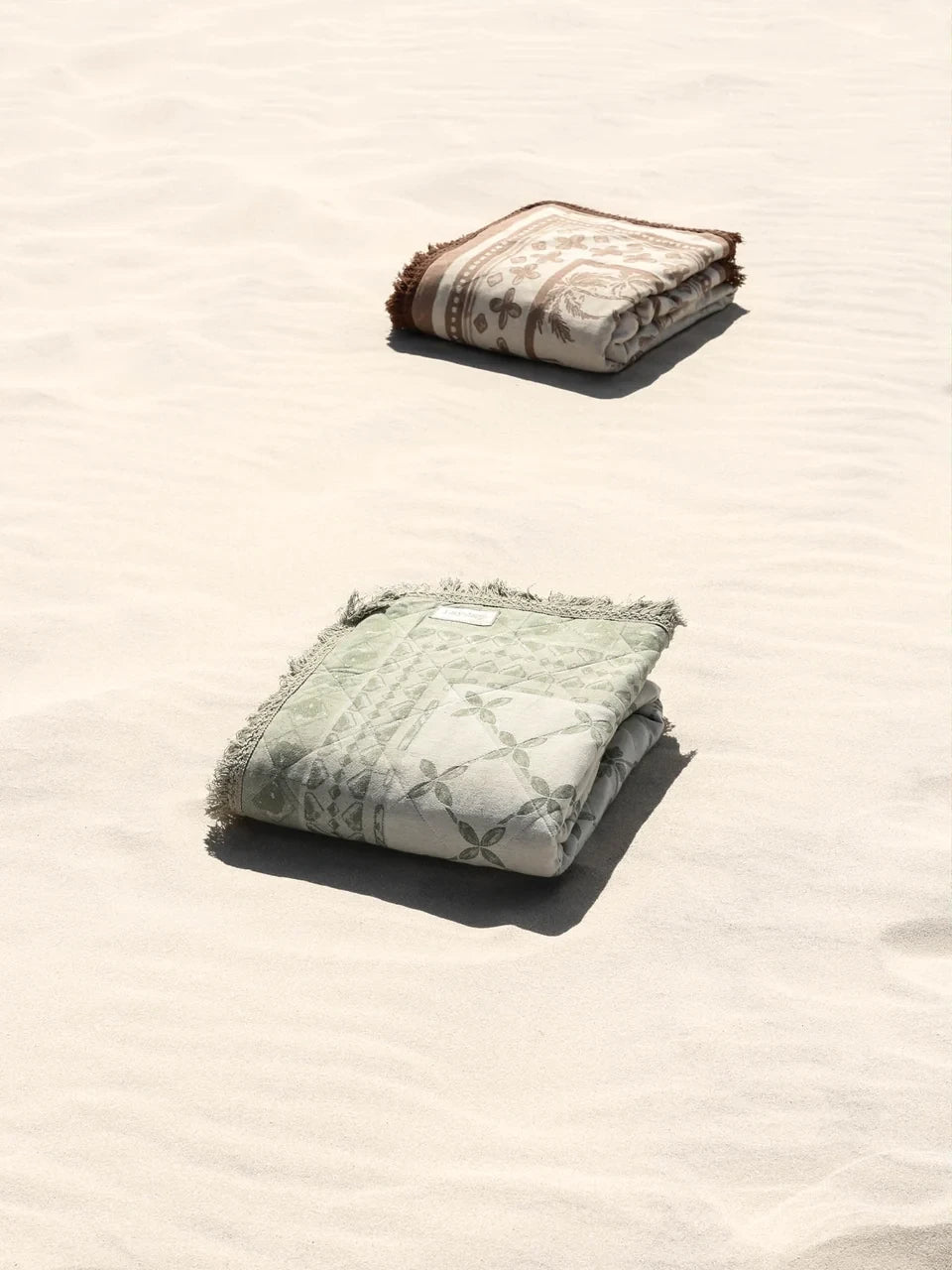 Two folded Layday blankets, one green and one patterned, on sandy beach.