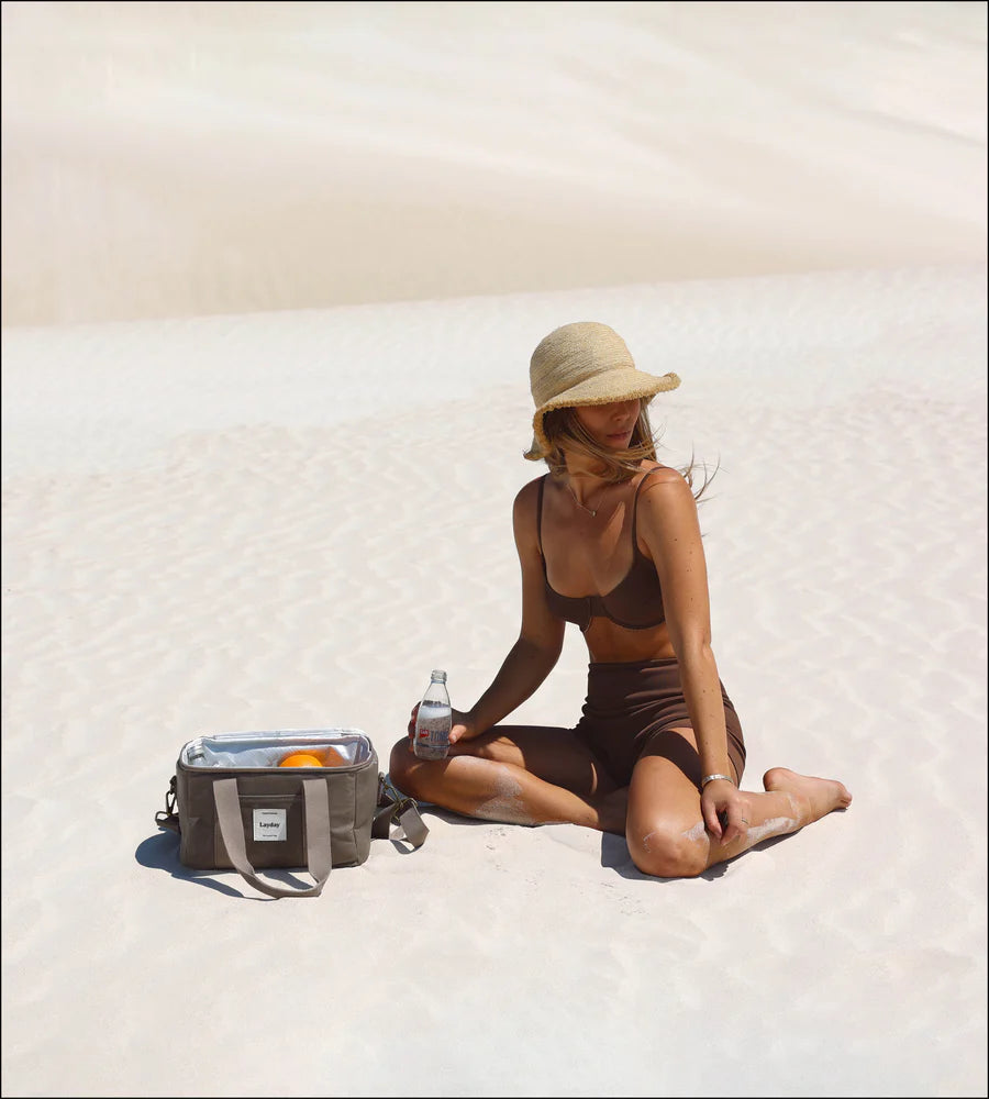 Woman sitting on a sandy beach with a Laydy cooler bag and bottle.