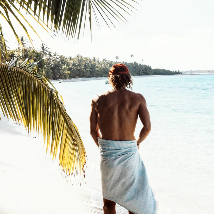 Man standing on a tropical beach with palm leaves and turquoise water.
