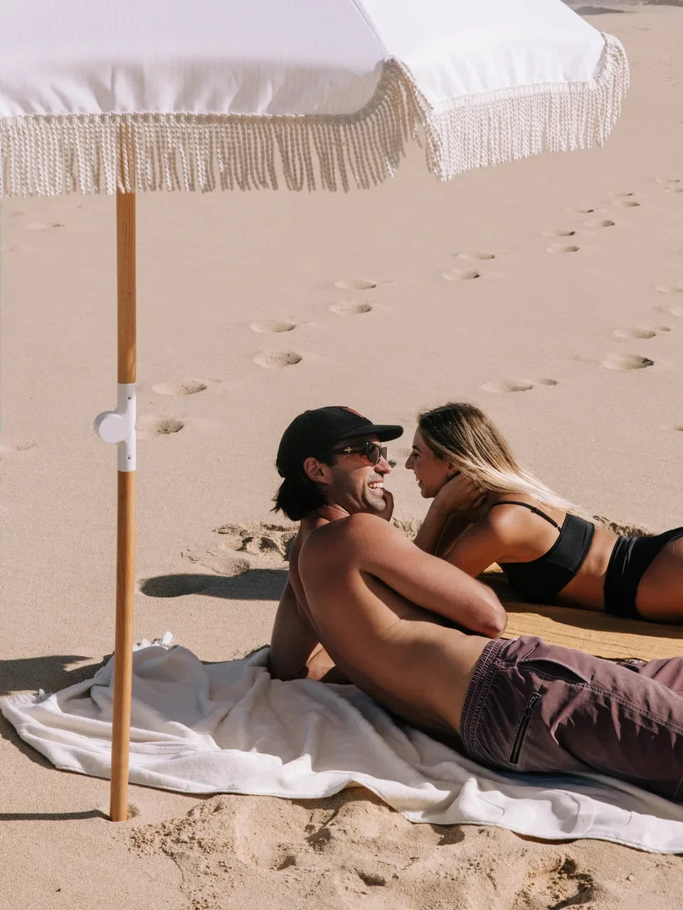 Two people lying on a Layday towel under a Layday umbrella on a sandy beach.